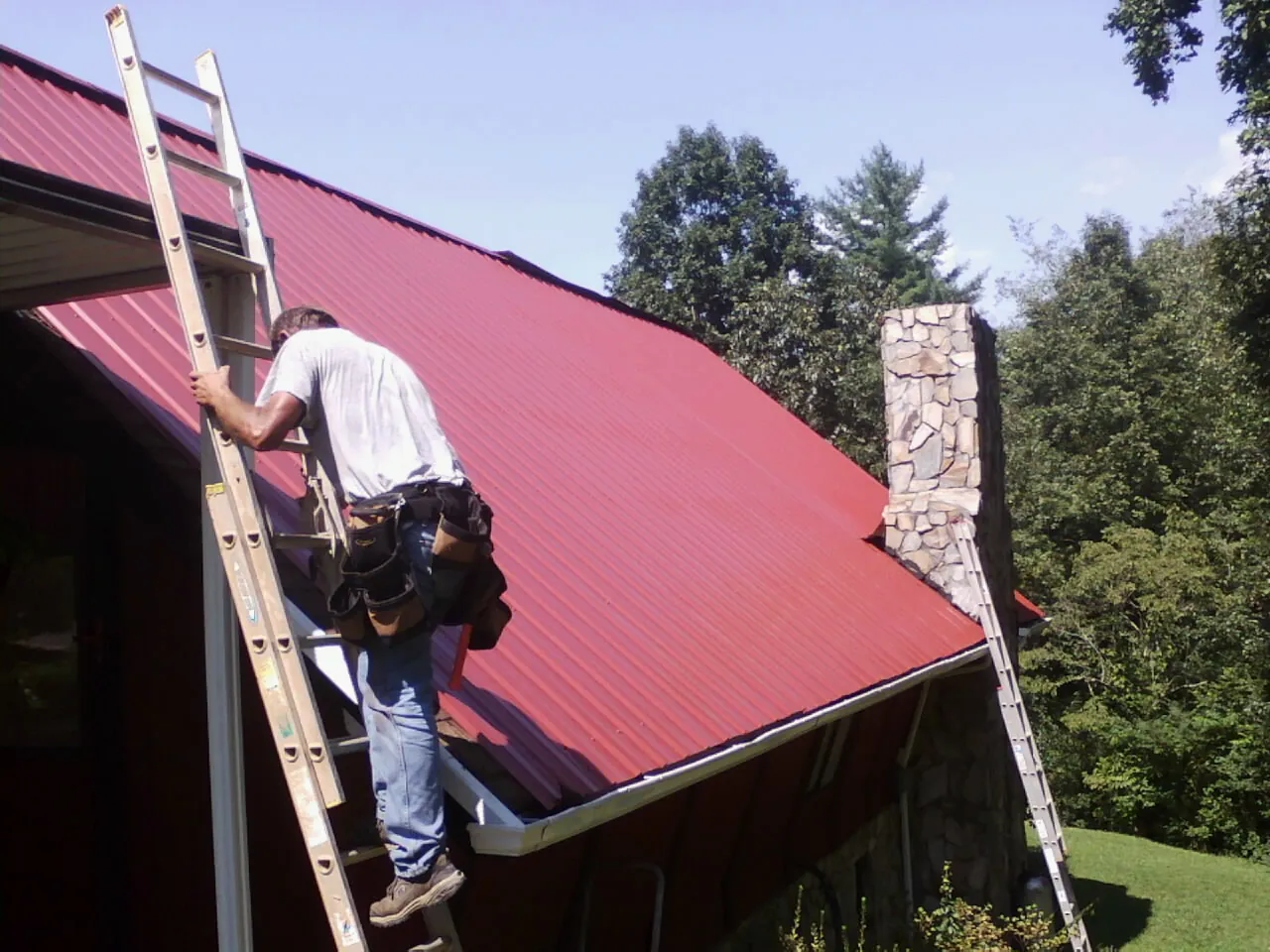 Roofer on a ladder beside a red metal roof and stone chimney — Soffit & Fascia Repair by Roofing Brothers in Northern Cambria